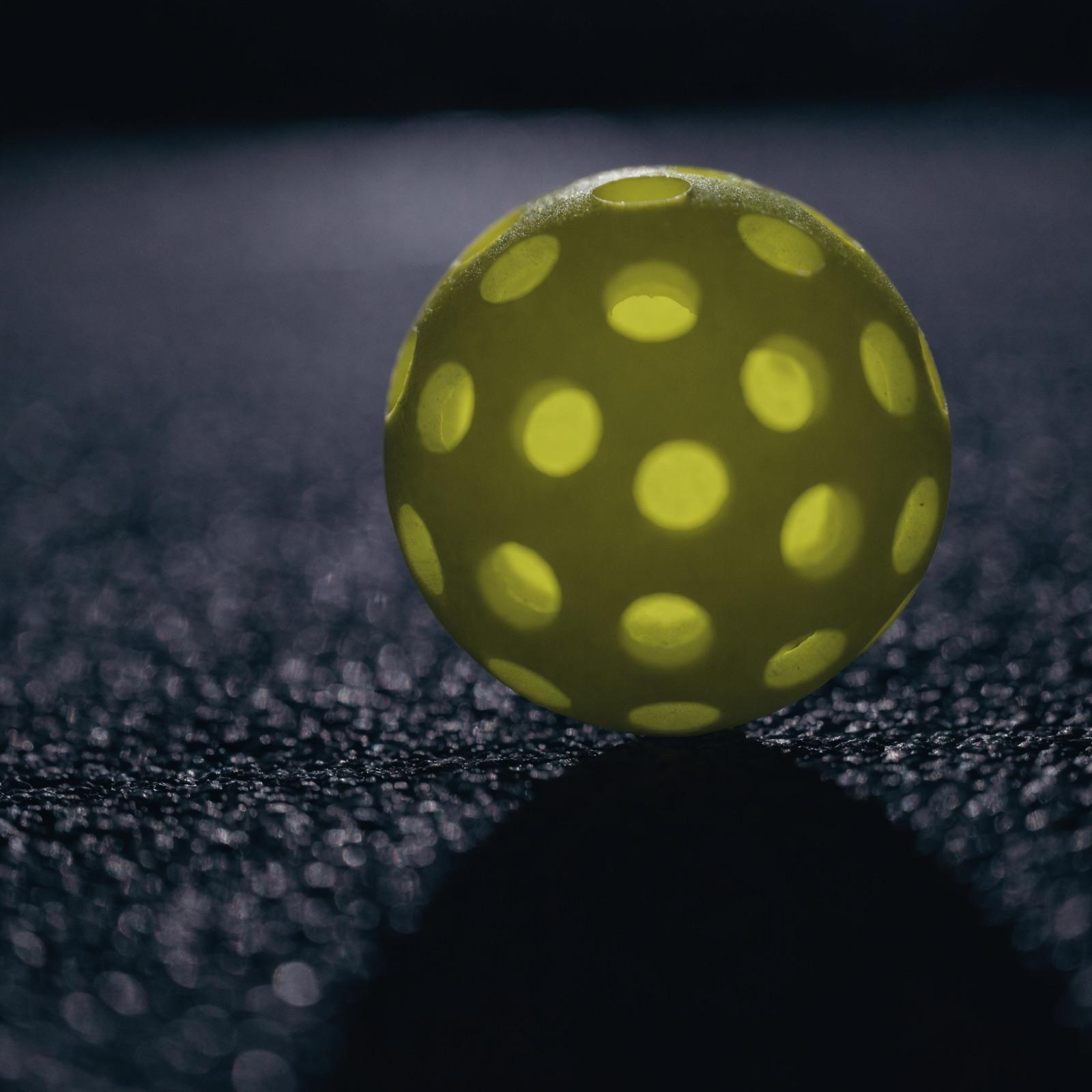 Macro close-up of a yellow perforated pickleball on textured court surface. Representative detail image.