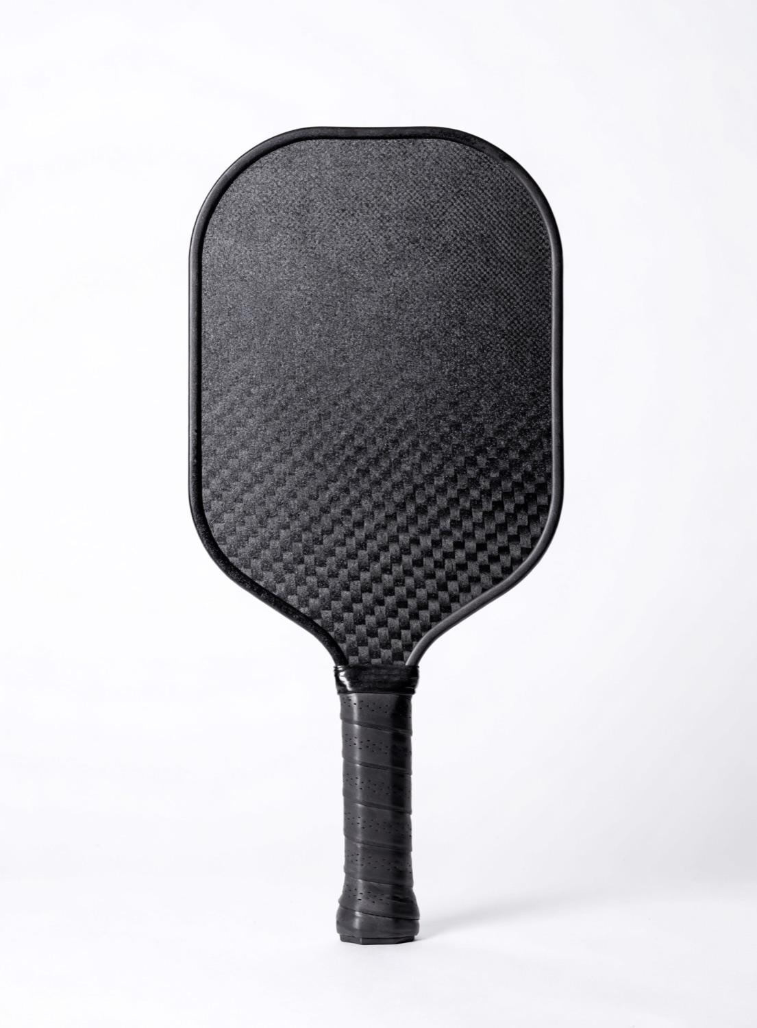 Representative photo of a textured raw-carbon pickleball paddle on a hard court; stands in for the Ronbus R1 Pulsar.